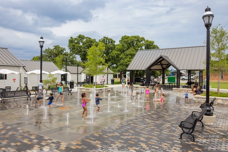 Families and children enjoying the interactive splash pad at Geyer Commons in Mansfield, Texas, with ground-level water jets, a central pavilion, and outdoor seating.