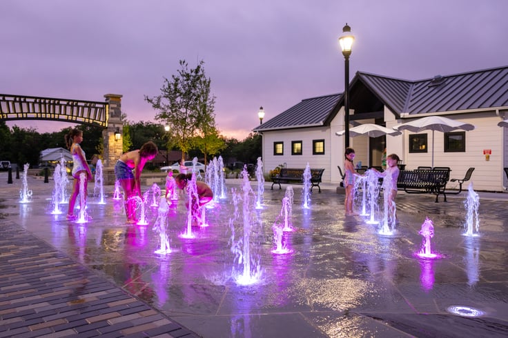 Families and children enjoying the interactive splash pad at Geyer Commons in Mansfield, Texas, with ground-level water jets, a central pavilion, and outdoor seating.
