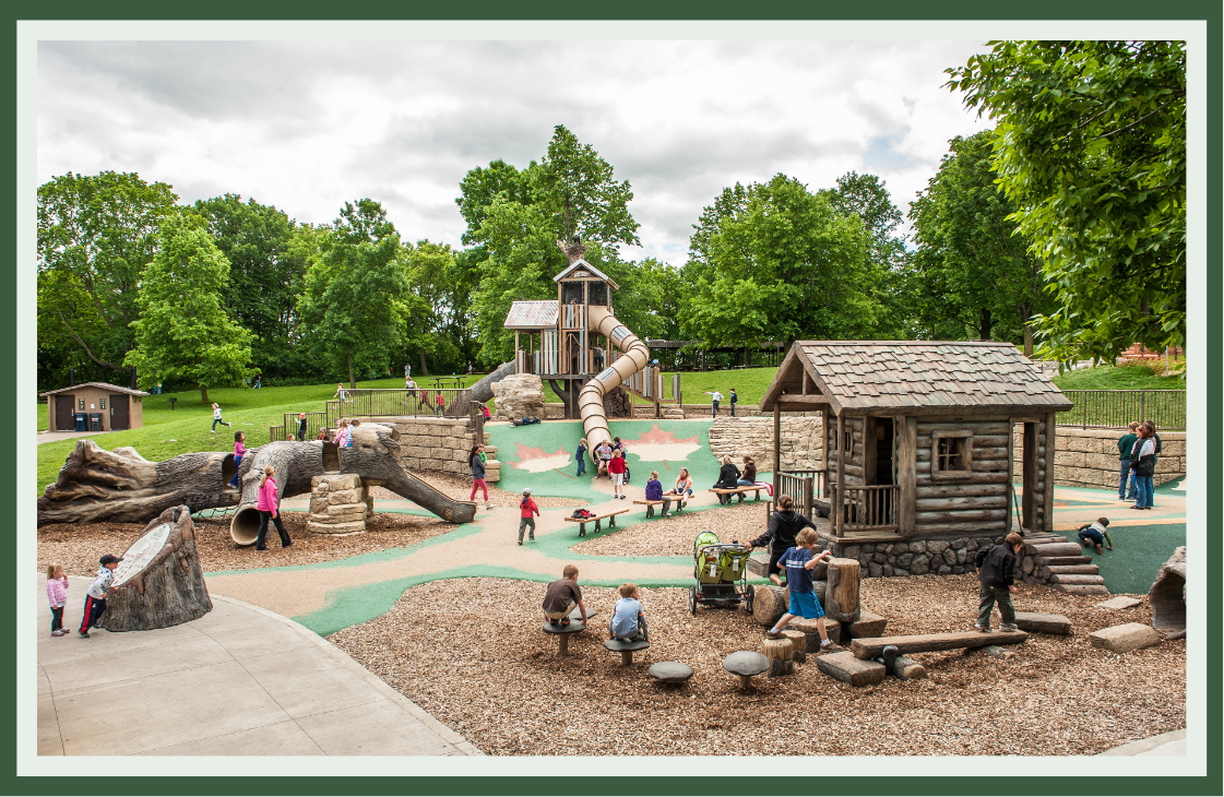 Nature-inspired playground with a log cabin playhouse, fallen-log climbers, tree-stump steppers and a treehouse-style tower with a tube slide, set among mature trees.
