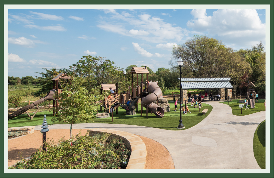 Wood-toned playground beside a curving accessible path, lamp posts and a shaded pavilion, with landscaping and seating for families.