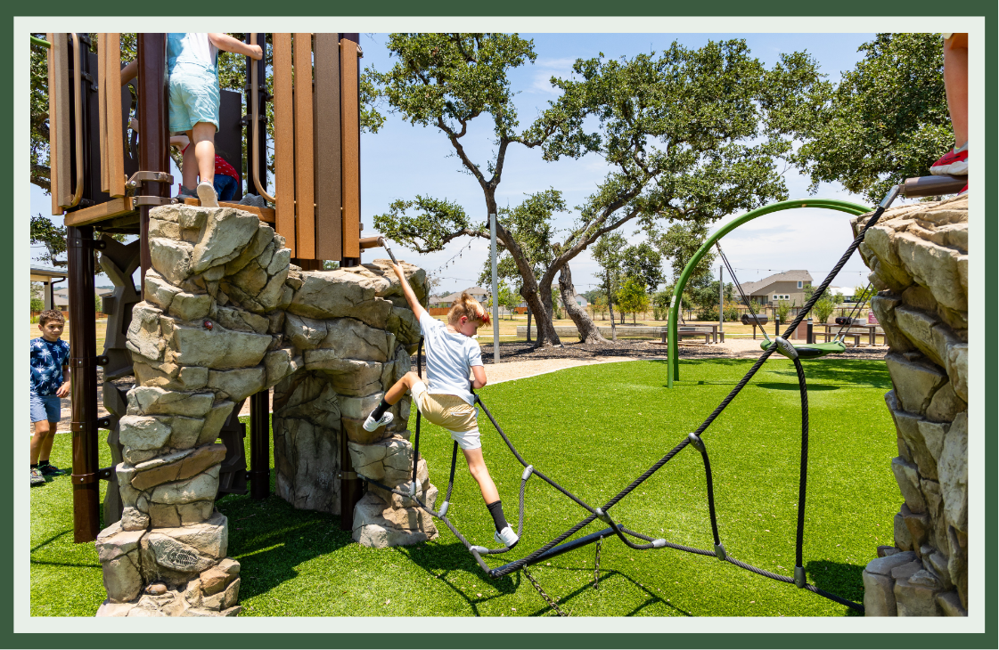 Child climbing a rope net between rock-textured climbing walls on a wood play tower, with a curved net climber and oak trees behind.