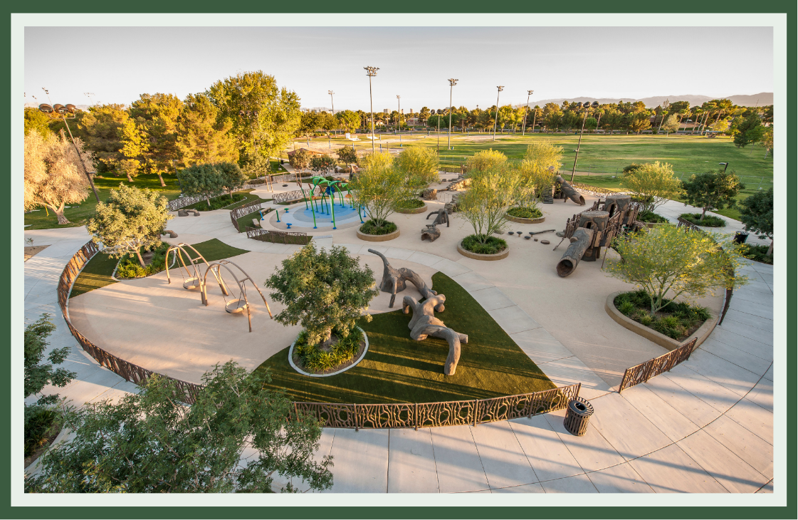 Aerial view of a circular playground with tree-filled planting islands, a lawn, climbable log and animal sculptures, and a splash pad nearby.