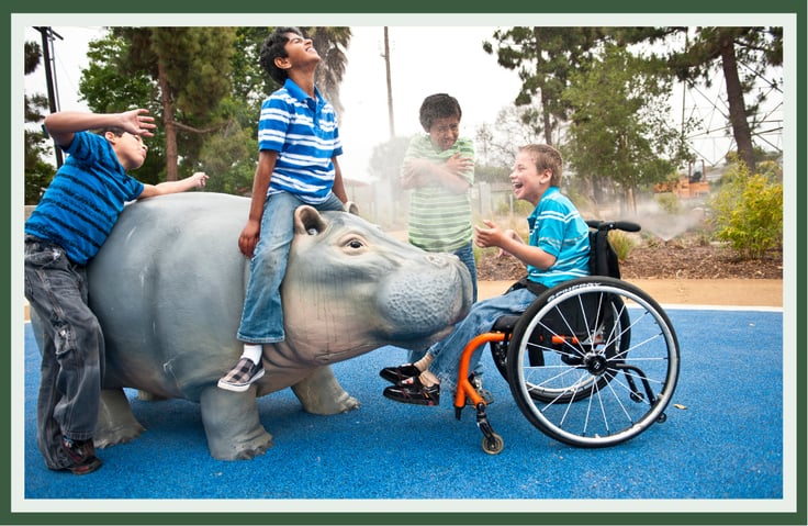 Children, including a child using a wheelchair, playing together on a life-size hippo sculpture on blue rubber surfacing.
