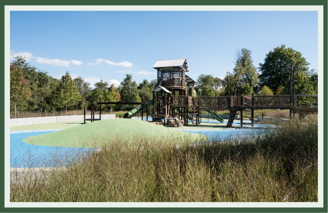 Treehouse-style playground on a grassy mound with a bridge crossing blue surfacing, surrounded by native grasses and mature trees.