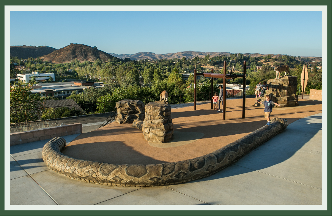 Rock-textured climbers and a serpentine boulder border on a hilltop play area, with sculpted forms echoing the rugged hills beyond.