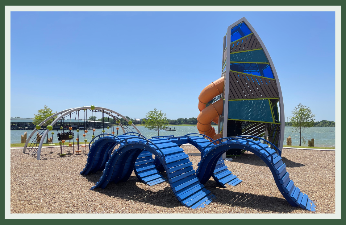 Blue wave-shaped climbers and a sail-shaped tower with an orange tube slide on a lakeside playground, with colors drawn from the water behind it.